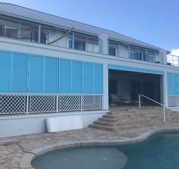 Custom light blue Bahama shutters installed on a luxury waterfront Florida home overlooking a pool, providing shade, privacy, and coastal elegance.