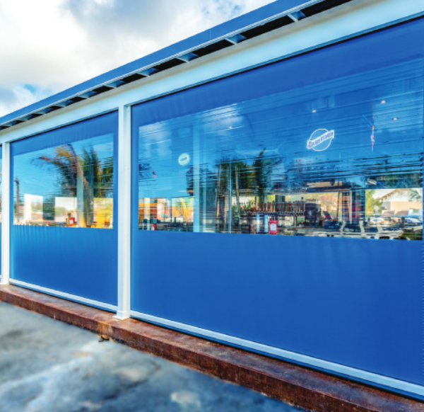 Blue outdoor enclosure curtains with clear vinyl windows installed on a commercial patio, providing weather protection while maintaining visibility and natural light.