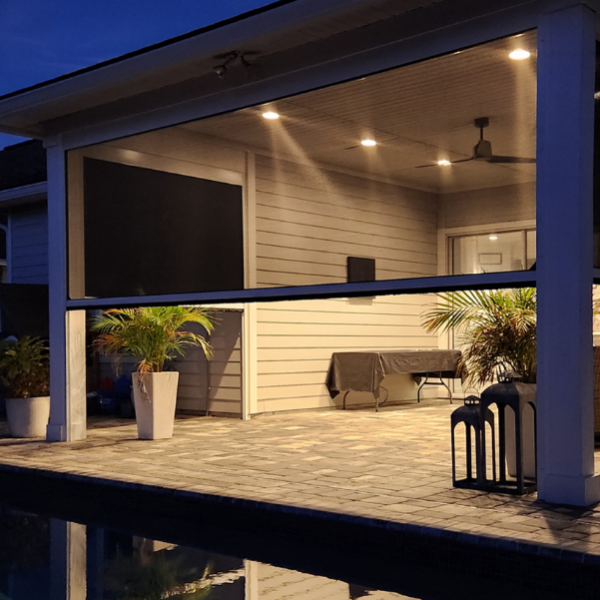 Motorized patio screen lowered on a covered outdoor living space at night, providing privacy, shade, and year-round comfort beside a pool.